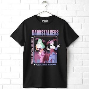 May include: A black short-sleeve t-shirt featuring a graphic print of the characters Morrigan and Lilith from Darkstalkers. The design includes the text DARKSTALKERS, MORRIGAN & LILITH, and VAMPIRE SAVIOR in a gradient blue and pink color scheme.