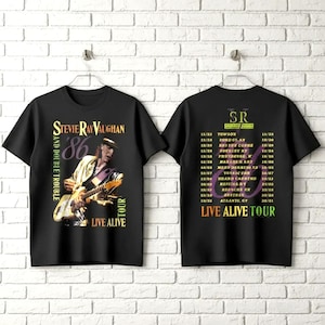 May include: Black t-shirts featuring a graphic of Stevie Ray Vaughan playing guitar, with "'86" and "LIVE ALIVE TOUR" text. The back of the shirt lists tour dates. The shirts are hanging on a white background.