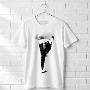 May include: A white short-sleeved t-shirt featuring a black and white graphic print of a person performing a high kick. The graphic shows the person in a dynamic pose with a shoe sole visible. The inner neck label reads American Classics.