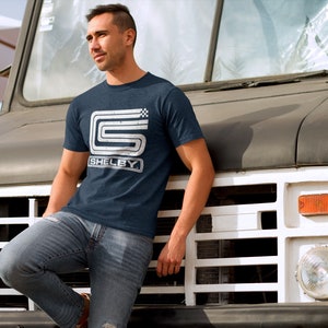 May include: A man wearing a navy blue t-shirt with a white vintage-style "Shelby" logo and a checkered flag design. He is standing in front of a white and black truck.