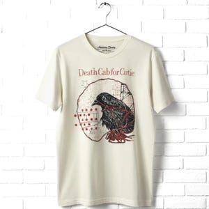 May include: A cream-colored short-sleeve t-shirt featuring a graphic print of a black bird entangled in red string. The text "Death Cab for Cutie" is printed in red above the bird, which is set against a circular background with stars and dots.