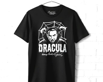 May include: A black short-sleeve t-shirt featuring a white graphic print of Dracula with a spiderweb background. The text reads DRACULA starring Bela Lugosi. The shirt has a crew neckline and is displayed on a hanger against a white brick wall.