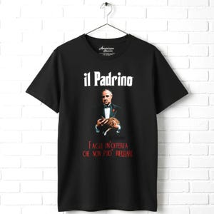 May include: Black t-shirt featuring a graphic of a man in a suit holding a cat, with the words "il Padrino" above and Italian text below. The shirt is hanging on a white brick wall.