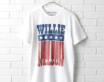 May include: A white short-sleeve t-shirt featuring a graphic print with the text WILLIE NELSON in blue and red block letters. The design includes a blue horizontal band with five white stars above red vertical stripes, all on a white fabric.