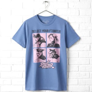 May include: A blue short-sleeve t-shirt featuring a graphic print with the text SELECT YOUR FIGHTER above four individual character portraits in pink squares. Below the portraits, the Street Fighter logo is printed in pink and white lettering.