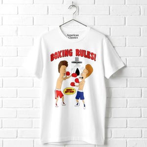 May include: A white short-sleeve t-shirt featuring a graphic of two cartoon characters boxing. The text "BOXING RULES!" appears in red above the characters. One character wears red shorts and the other blue shorts, both wearing red boxing gloves.
