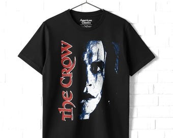 May include: A black short-sleeve t-shirt featuring a graphic print of a face with white and blue makeup. The text "the crow" is printed vertically in red on the left side of the graphic. The shirt has a crew neckline and is displayed on a hanger.