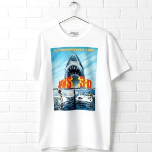 May include: A white short-sleeve t-shirt featuring a graphic print of the Jaws 3-D movie poster. The design displays a large shark with an open mouth, the text "THE THIRD DIMENSION IS TERROR" at the top, and "JAWS 3-D" in large yellow letters.