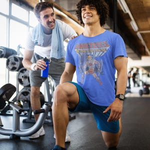 May include: A blue t-shirt with the text "MASTERS OF THE UNIVERSE" and a graphic of a cartoon character. The person is wearing blue shorts and a black watch. Another person is standing behind him with a white towel and a blue water bottle.