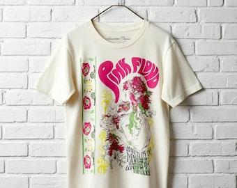 Puede incluir: Camiseta color crema con un gráfico de Pink Floyd. El diseño incluye el nombre de la banda en rosa, una ilustración floral y el texto "MARCH 11-15 66 LONDON WOOD". La camiseta está colgada en una percha negra contra una pared de ladrillos blancos.