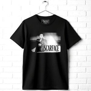 May include: A black short-sleeve t-shirt featuring a black and white graphic print of a man holding a firearm. The text SCARFACE is printed in white below the image. The inner neck label displays the text American Classics.