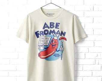 Puede incluir: Una camiseta de manga corta color crema con un gráfico de una salchicha de dibujos animados que lleva una corona. El texto dice ABE FROMAN SAUSAGE KING OF CHICAGO y FERRIS BUELLER'S DAY OFF. El diseño incluye un círculo azul y un tenedor.