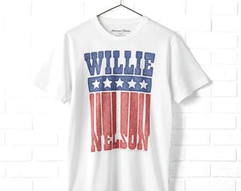 May include: A white short-sleeve t-shirt featuring a graphic print with the text WILLIE in blue, a blue horizontal band with five white stars, and the text NELSON in red vertical stripes. The design has a textured, glittery appearance.