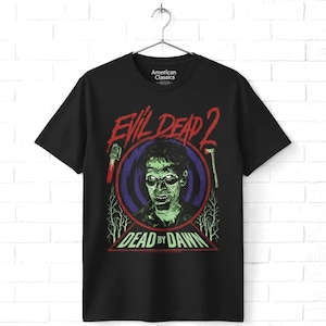 May include: A black short-sleeve t-shirt featuring a graphic print from the movie Evil Dead 2. The design includes a central portrait of a character, the text Evil Dead 2 in red, and Dead by Dawn in green, with a chainsaw and axe illustration.