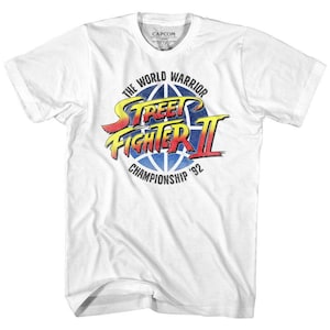 May include: White t-shirt featuring the Street Fighter II logo. The design includes the text "THE WORLD WARRIOR" above a blue globe with "STREET FIGHTER II" in yellow and red, and "CHAMPIONSHIP '92" below. A Capcom label is visible.