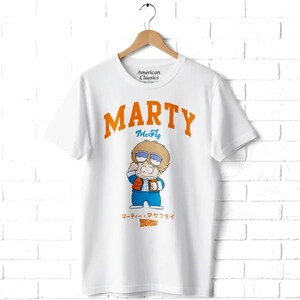 May include: A white short-sleeve t-shirt featuring the orange text MARTY and blue text McFly above a cartoon illustration of a character wearing sunglasses, a red vest, and blue jacket. Below the character is Japanese text and the Back to the Future logo.