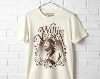 May include: A cream-colored short-sleeve t-shirt featuring a brown graphic print of Willie Nelson in profile. The design includes the text Willie Nelson and Tour '77 inside an ornate circular frame. The inner neck label reads American Classics.