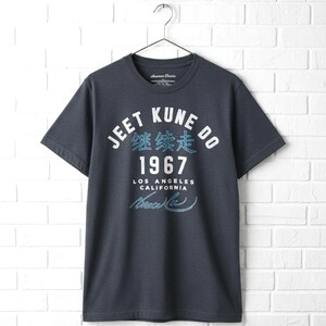 May include: A dark charcoal gray short-sleeve t-shirt featuring white and light blue text that reads JEET KUNE DO, 1967, LOS ANGELES CALIFORNIA, and a signature. The shirt includes Chinese characters and is displayed on a metal hanger.