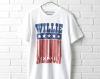 May include: White t-shirt with a patriotic design featuring the name "WILLIE NELSON" in blue and red glitter text. The design includes blue stars and red stripes, reminiscent of the American flag. The shirt is hanging on a metal hanger.