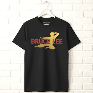 May include: A black short-sleeve t-shirt featuring a gold silhouette of a person in a martial arts pose. The text BRUCE LEE is printed in red across the center of the graphic. The shirt is displayed on a white hanger against a white brick wall.