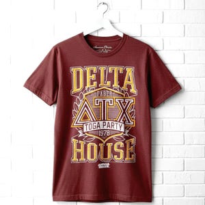 May include: A maroon short-sleeve t-shirt featuring a graphic print that reads DELTA HOUSE, FABER, ATX, TOGA PARTY, 1978, and ANIMAL HOUSE. The design uses yellow, white, and maroon lettering with a laurel wreath and triangular Greek letter symbol.