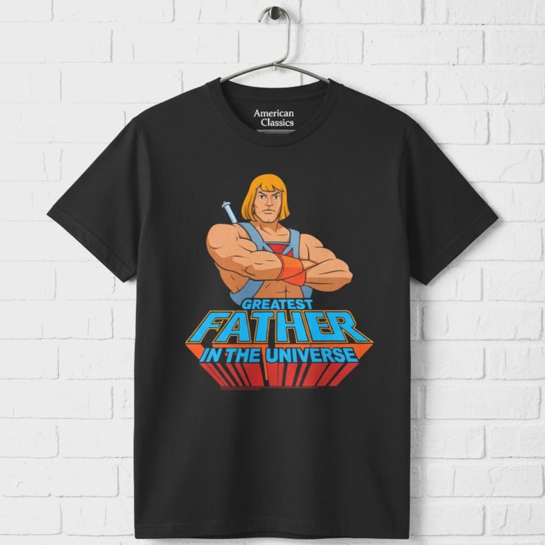 May include: Black t-shirt featuring a cartoon illustration of a muscular blond man with a sword, and the text "GREATEST FATHER IN THE UNIVERSE" in blue and red. The shirt has a crew neck and the brand "American Classics" is printed on the upper left.