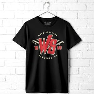May include: Black t-shirt with a red and white graphic. The graphic features the text "WYLD STALLYNS" above a stylized "WS" logo, with "1989" and "SAN DIMAS, CA" below. The shirt is hanging on a metal hanger.