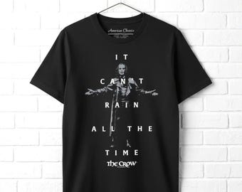 May include: A black short-sleeve t-shirt featuring a graphic of a person with arms outstretched. The white text reads IT CAN'T RAIN ALL THE TIME and THE CROW. The shirt has a crew neckline and is displayed on a hanger against a white brick wall.