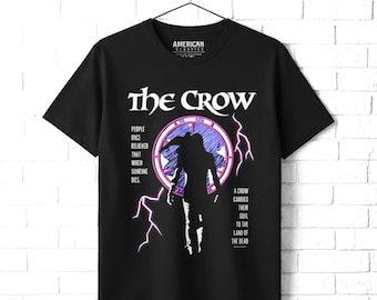 May include: A black short-sleeve t-shirt featuring a graphic of The Crow. The design includes a silhouette of a person, a circular purple frame, lightning bolts, and the text: People once believed that when someone dies, a crow carries their soul to the land of the dead.