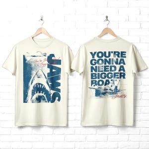 May include: An off-white short-sleeve t-shirt featuring a blue graphic of a shark and the text JAWS on the front, and the text YOU'RE GONNA NEED A BIGGER BOAT with a boat illustration on the back. The design includes red script accents.