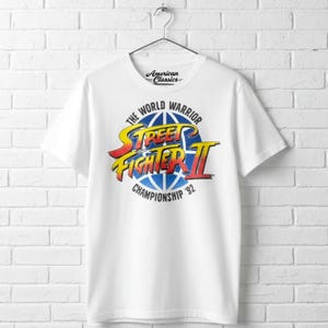 May include: A white short-sleeve t-shirt featuring a graphic print on the chest. The design includes the text Street Fighter II in yellow and red, with the words The World Warrior Championship '92 and an American Classics logo above.