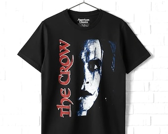 May include: A black short-sleeve t-shirt featuring a graphic print of a face with white makeup and the red text "the crow" vertically aligned on the left side. The inner neck label displays the text "American Classics".