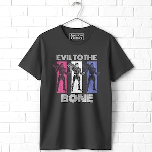 May include: A black short-sleeve t-shirt featuring a graphic print with the text "American Classics" at the neckline. The center design displays three skeletal figures in pink, white, and blue vertical panels with the text "EVIL TO THE BONE".