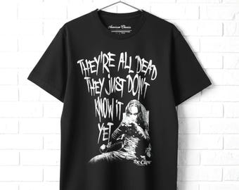 May include: A black short-sleeve t-shirt featuring a white graphic print of a character from The Crow. The text reads, "THEY'RE ALL DEAD THEY JUST DON'T KNOW IT YET." The shirt has a crew neckline and is displayed on a metal hanger against a white wall.