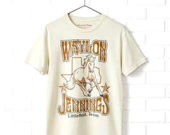 May include: A cream-colored short-sleeve t-shirt featuring a graphic of a horse centered over a Texas state outline. The text reads Waylon Jennings and Littlefield, Texas in a distressed brown font with two stars on the sides.