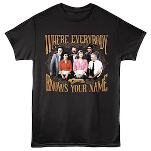 May include: Black t-shirt featuring a group of people in a framed design with the text "WHERE EVERYBODY KNOWS YOUR NAME". The design is in shades of brown and gold. The word "Cheers" is also visible.