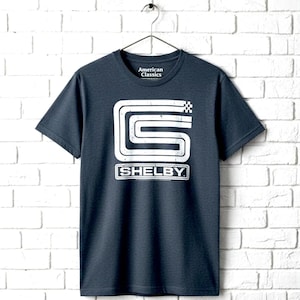 May include: A navy blue t-shirt with a white graphic of the word "Shelby" in a rectangular box, and a stylized "S" with a checkered flag design. The shirt is hanging on a metal hanger against a white brick wall. The text "American Classics" is printed on the shirt.