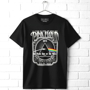 May include: A black short-sleeve t-shirt featuring a graphic print of the Pink Floyd The Dark Side of the Moon 1973 tour. The design includes a white prism, a rainbow spectrum, and text reading An Evening of Sight and Sound with Pink Floyd.