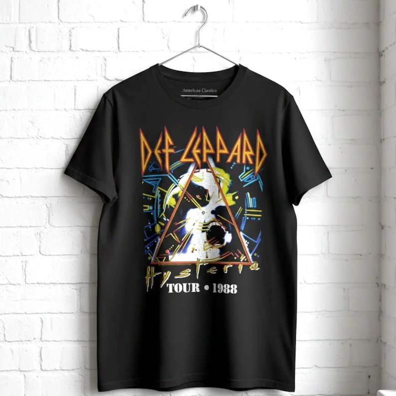May include: A black short-sleeve t-shirt featuring a graphic print of the Def Leppard Hysteria Tour 1988. The design includes a central triangle, abstract blue and yellow geometric lines, and the band name in orange lettering on a black background.