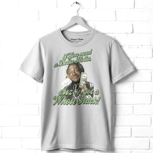 May include: A light gray short-sleeved t-shirt featuring a graphic of a person holding a dollar bill. The text reads: if You need a Dolla Holla cuz I got a Whole Stack! The shirt is displayed on a gray hanger against a white brick wall background.