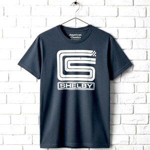 May include: A dark charcoal grey short-sleeve t-shirt featuring a white distressed graphic of a stylized letter S and a checkered flag icon above the text SHELBY. The inner neck label displays the text American Classics in white lettering.