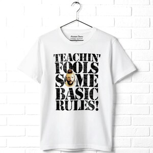 May include: A white short-sleeve t-shirt featuring a distressed black graphic print that reads TEACHIN' FOOLS SOME BASIC RULES! with a small photographic portrait of a man with a mohawk hairstyle positioned in the center of the text.