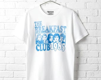 May include: A white short-sleeved t-shirt featuring a blue graphic print of the five main characters from the movie The Breakfast Club. The text reads The Breakfast Club 1985 in a distressed blue font. The shirt has a crew neckline and short sleeves.