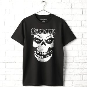 May include: Black t-shirt with a white graphic of a skull and the word "SKELETOR" above it. The shirt is hanging on a metal hanger against a white brick wall. The shirt has a crew neck and short sleeves.