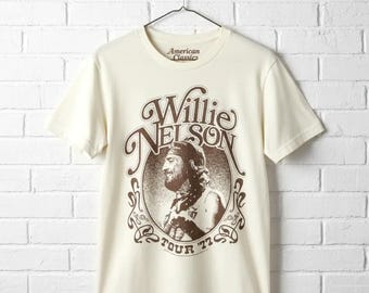 May include: Cream-colored t-shirt featuring a brown graphic of Willie Nelson with the text "Willie Nelson" and "Tour '77". The shirt is hanging on a metal hanger against a white brick wall. The text "American Classics" is printed on the shirt.
