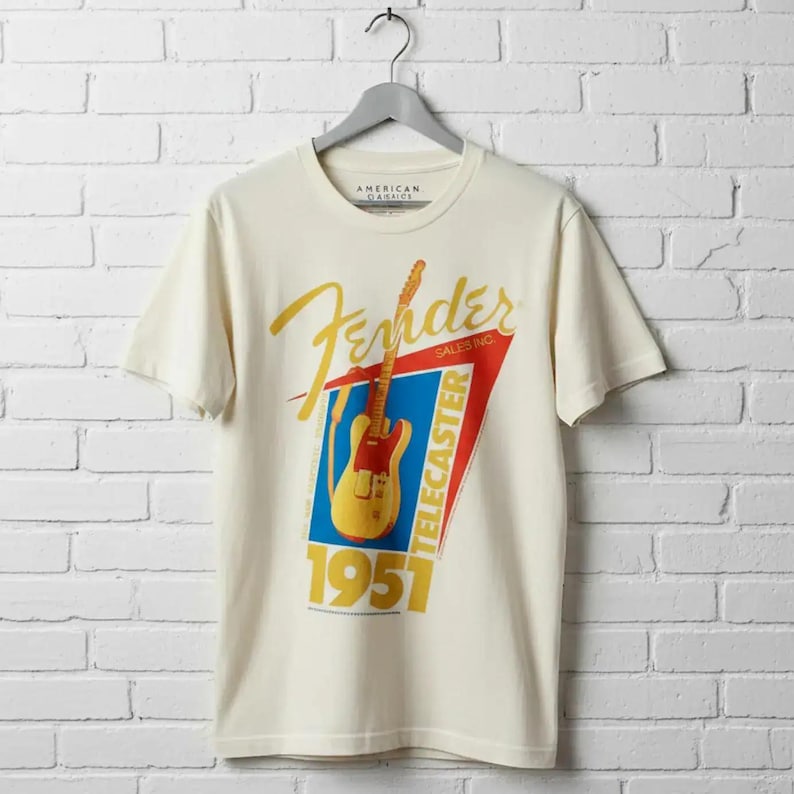 May include: Cream-colored t-shirt featuring a Fender Telecaster guitar graphic. The design includes the Fender logo in gold script, a red and blue triangle, and the text "TELECASTER 1951". The shirt is hanging on a gray hanger against a white brick wall.