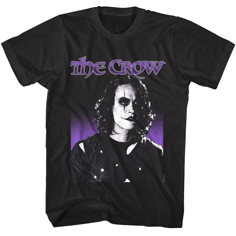 May include: Black t-shirt featuring a portrait of a person with white face makeup and dark eye makeup, with the text "The Crow" in purple above the portrait. The background is purple.