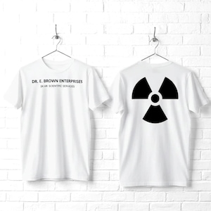 May include: A white short-sleeved t-shirt featuring the black text DR. E. BROWN ENTERPRISES 24 HR SCIENTPIC SERVICES on the front and a large black radiation hazard symbol printed on the back, both displayed on hangers.