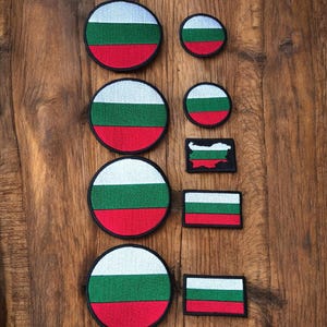 May include: A selection of embroidered patches displaying the Bulgarian flag. The patches are in diverse shapes and sizes, including circles, rectangles, and a map outline. The flag design features white, green, and red horizontal stripes.