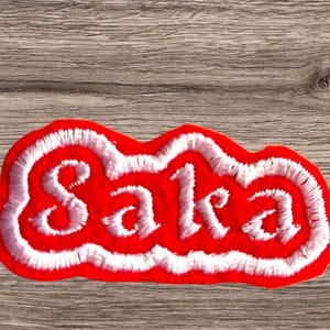 May include: Red felt patch with white embroidered lettering that reads "Saka".